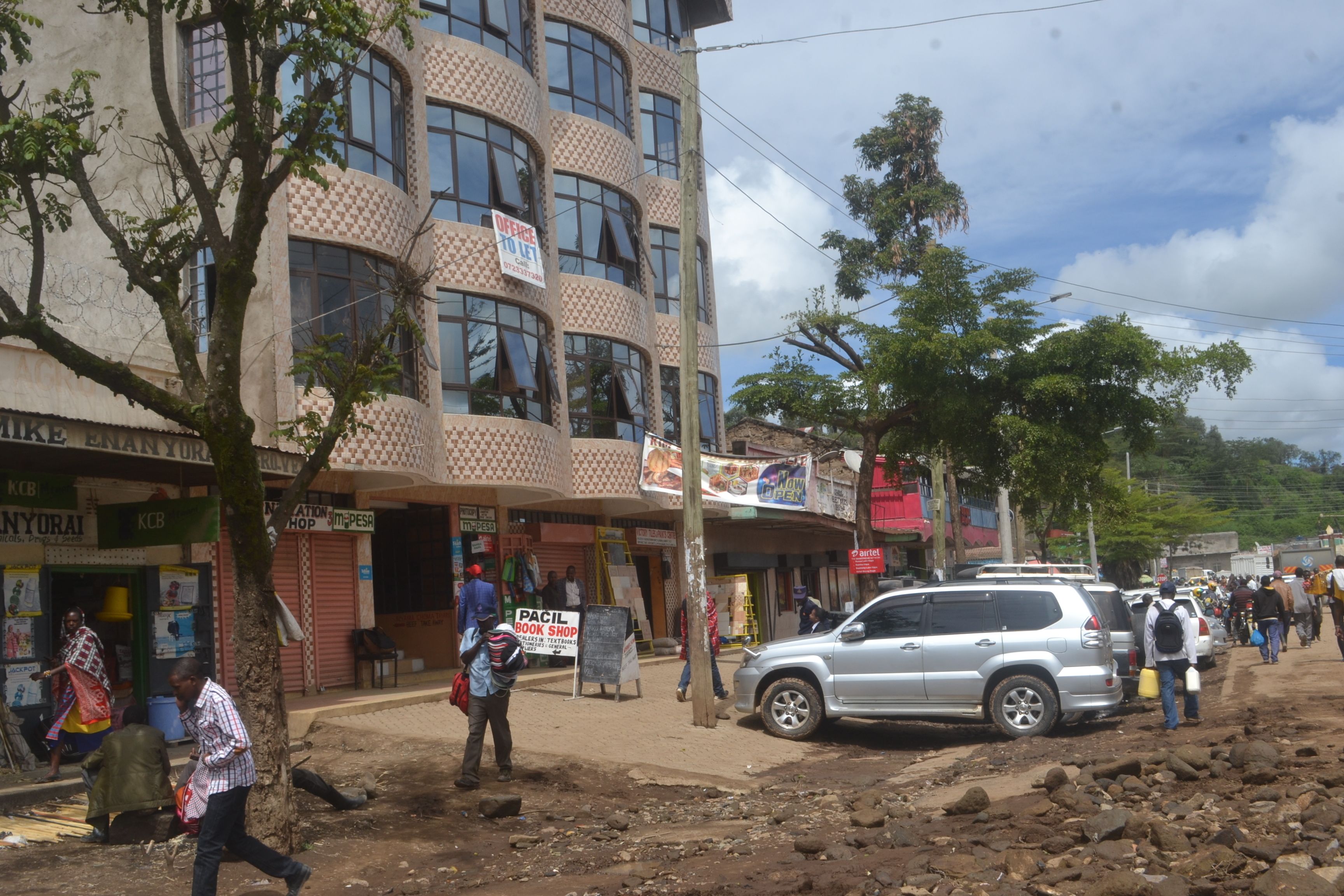 A multi-story building with a checkered facade, a visible ground floor with shops, and a parked car on a dirt road.