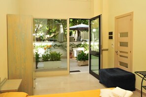 Habitación con puerta corrediza de vidrio que da a un espacio exterior con plantas y una mesa. Hay un banco y una puerta con cerradura de teclado.