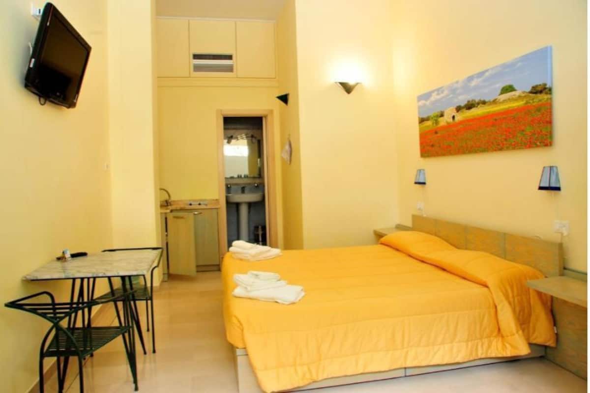 Habitación de hotel con cama, una mesita, una silla, un televisor y un cuadro en la pared.