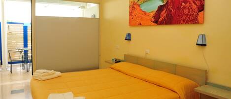 A hotel room with a large bed, a painting of a natural landscape, a desk with a lamp, and a view of the outside.