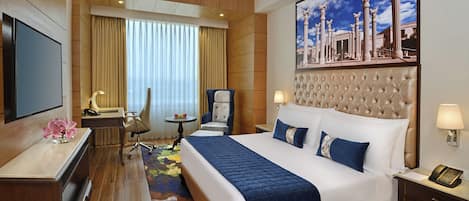 A hotel room with a large bed, a desk, a chair, a small table, and a television.
