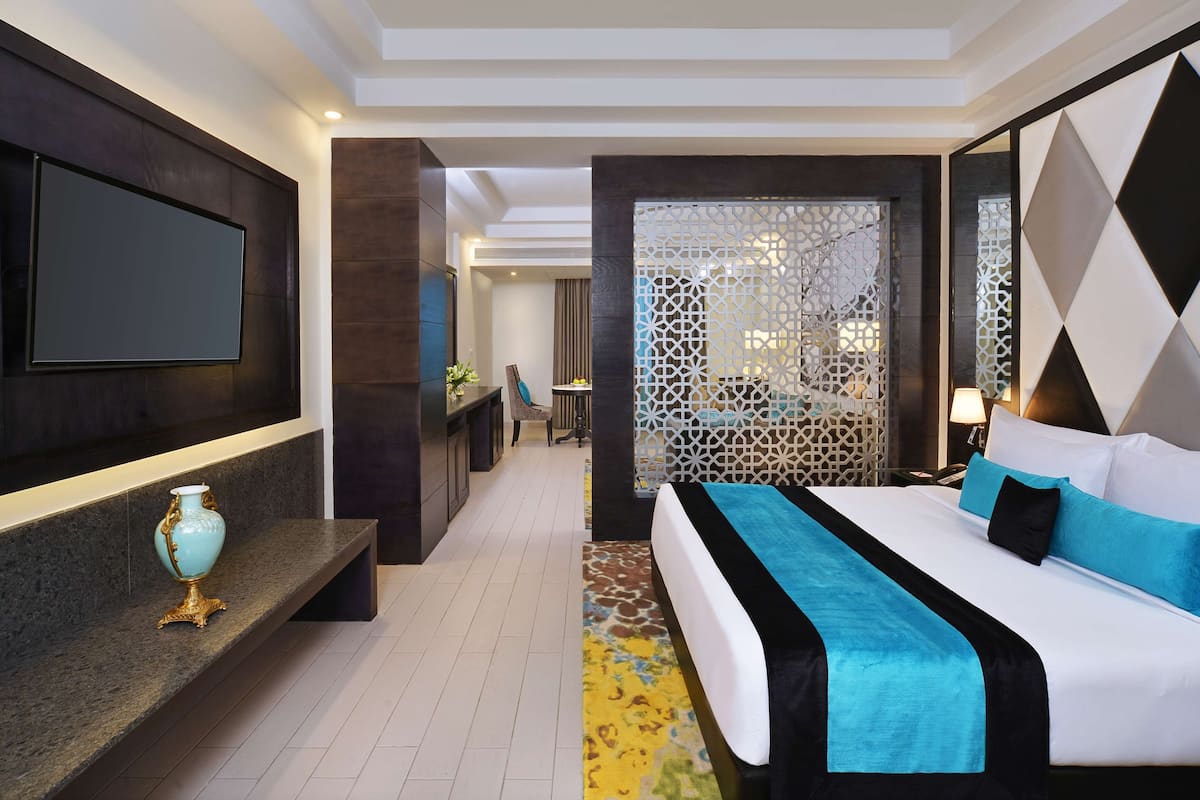 Une chambre d’hôtel moderne dotée d’un grand lit, d’un téléviseur à écran plat fixé au mur, d’un petit coin salon avec une chaise et une table, et d’un paravent décoratif aux motifs géométriques.