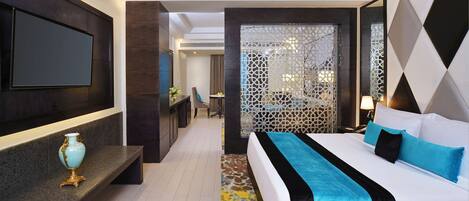 A modern hotel room with a large bed, a flat-screen TV mounted on the wall, a small seating area with a chair and table, and a decorative screen with geometric patterns.