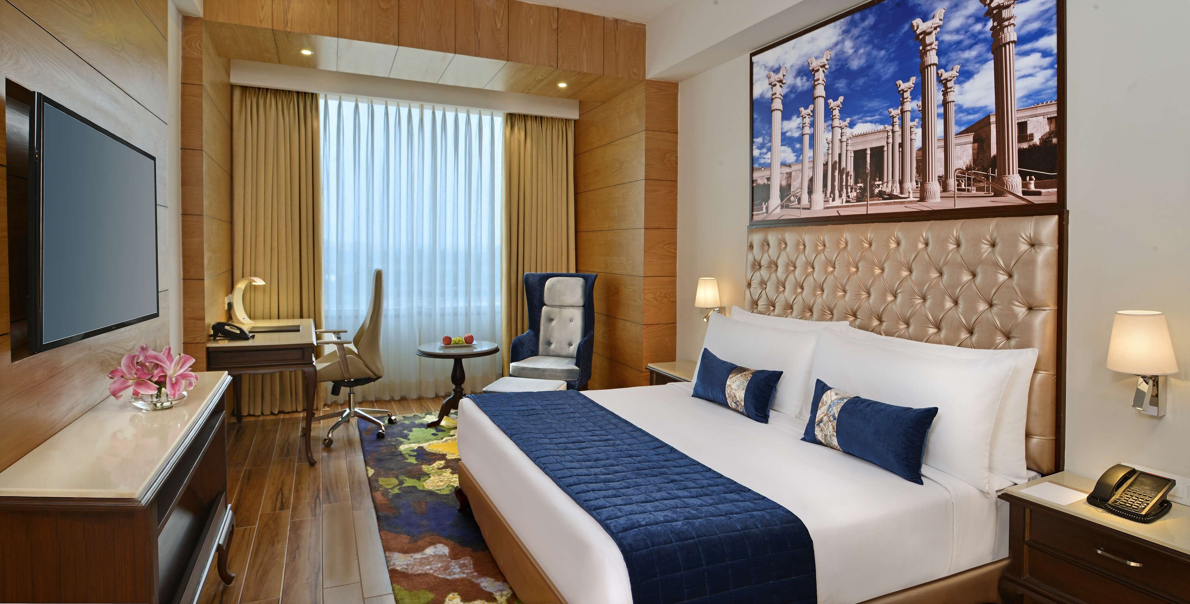 Une chambre d’hôtel avec un grand lit, un bureau avec un téléphone, une chaise, une petite table avec une lampe et un tableau représentant des colonnes sur le mur.