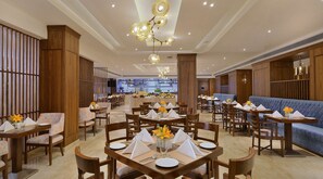 Uma ampla área de refeições com várias mesas preparadas para o serviço, um espaço de estar central com um sofá azul, e uma cozinha visível ao fundo.