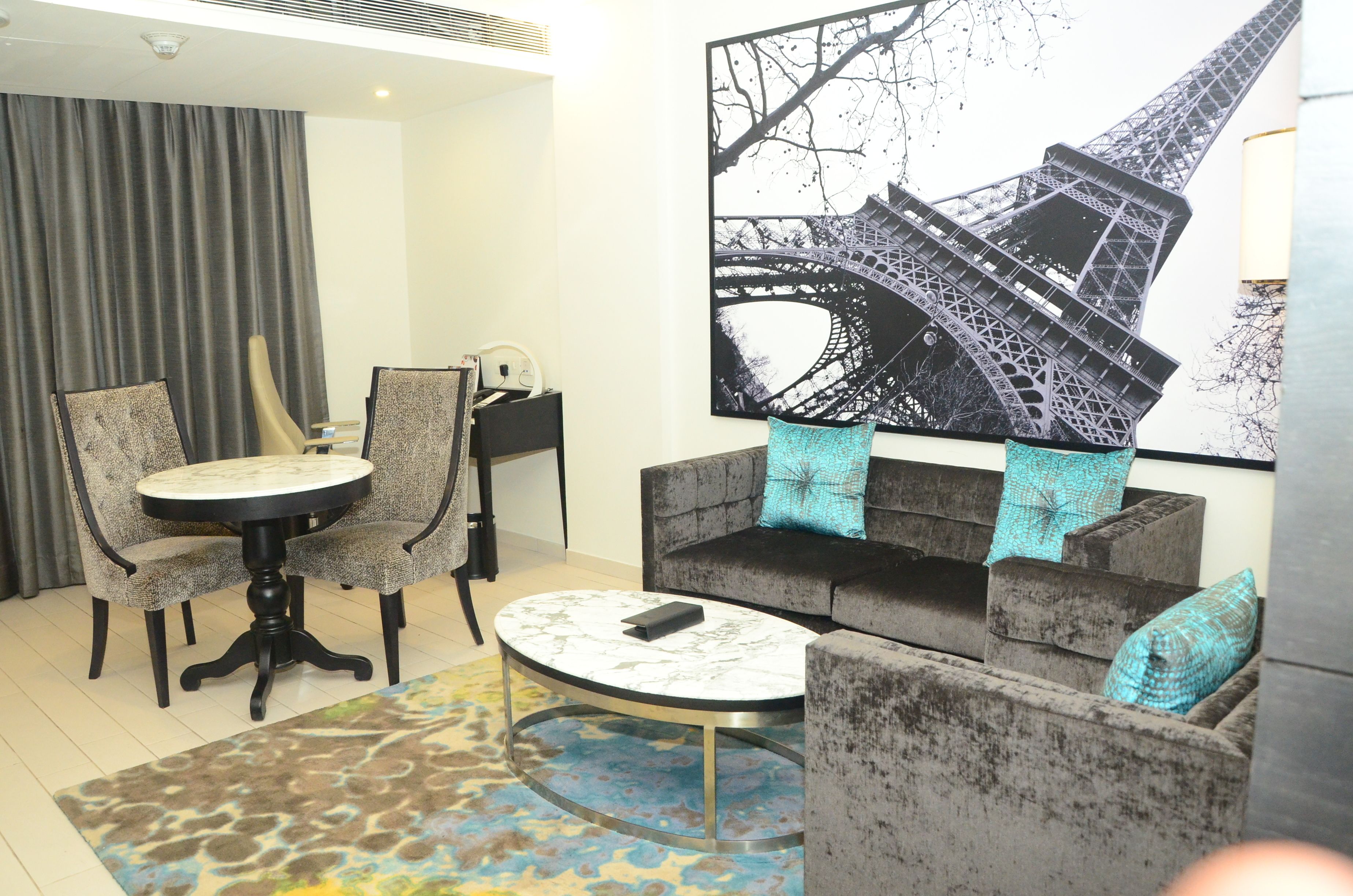 A hotel room with a round table, two armchairs, a sofa, and a large Eiffel Tower artwork on the wall.