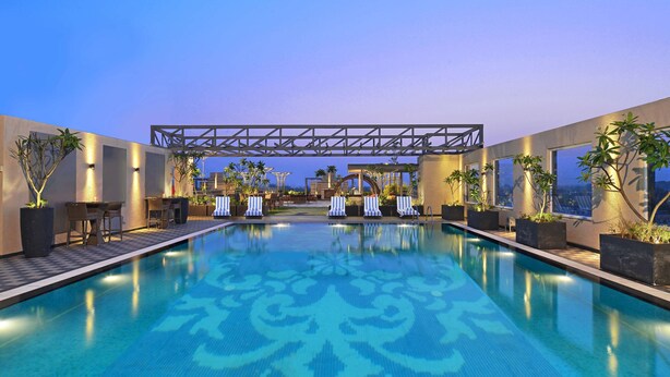 A swimming pool with a geometric pattern, surrounded by lounge chairs and potted plants, with a view of the city skyline at dusk. - Radisson Chandigarh Zirakpur (Dera Bassi)