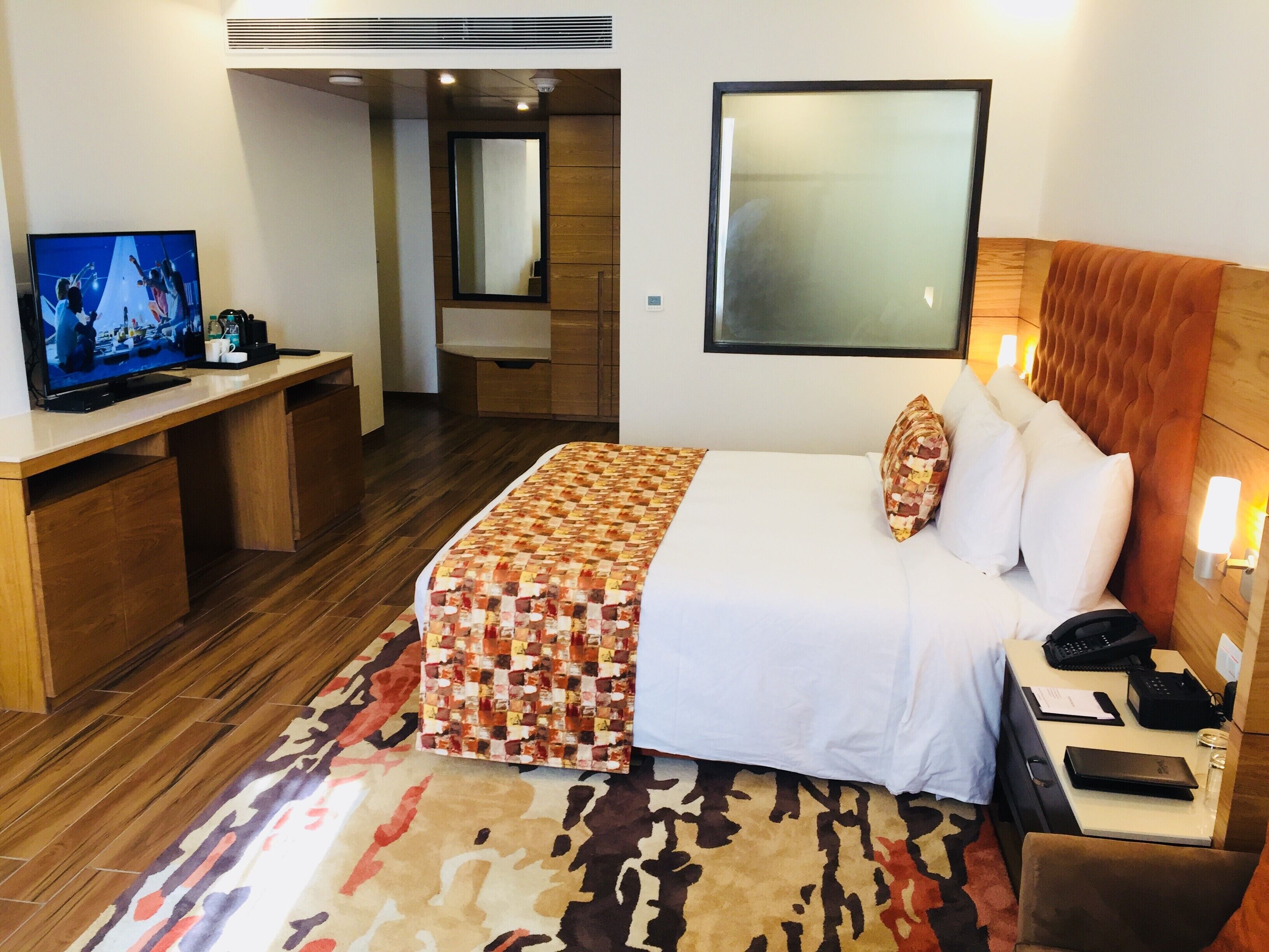 Habitación de hotel con cama, escritorio con televisor, espejo y lámpara de pared.