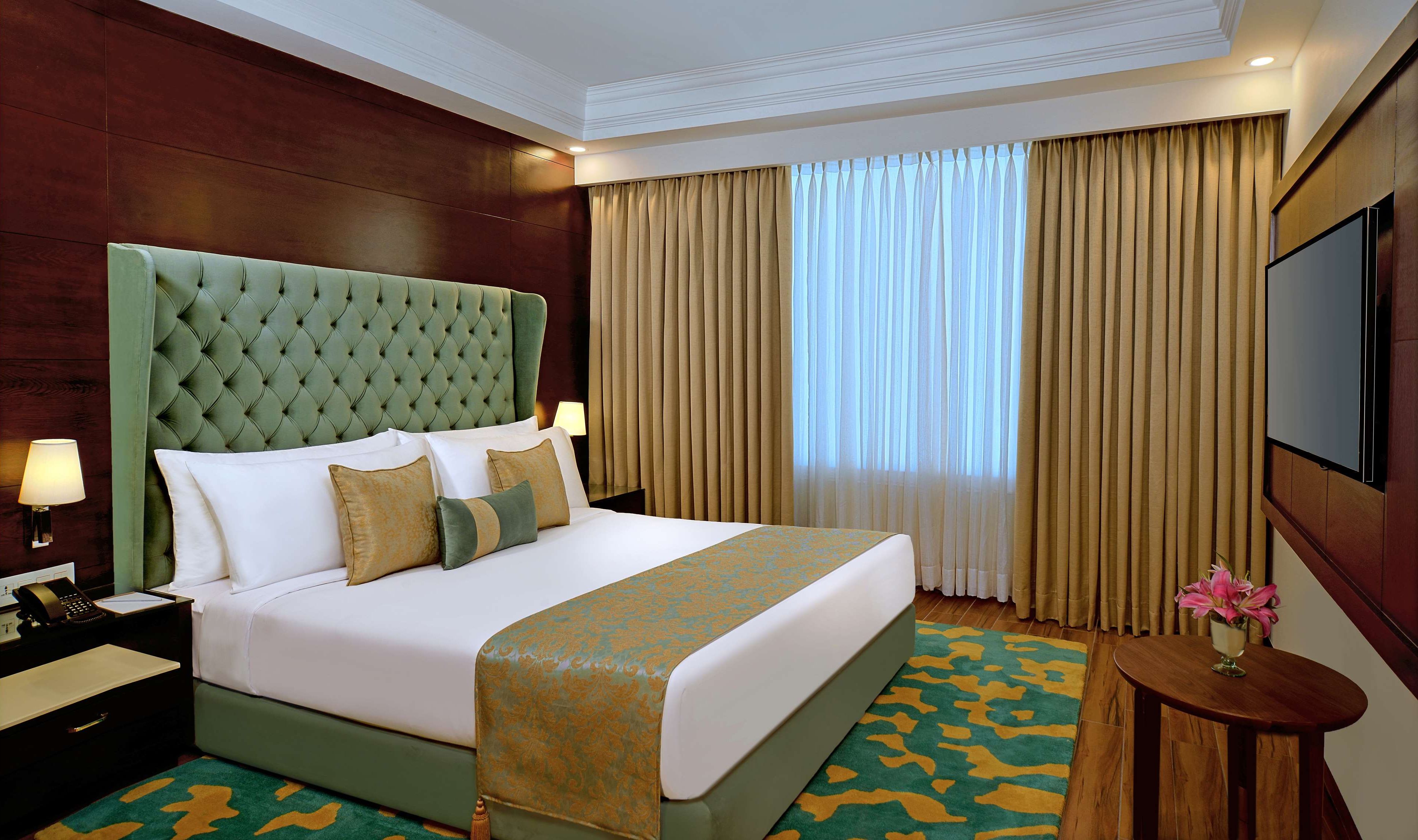 A hotel room with a large bed, a green upholstered headboard, a wooden nightstand with a lamp, a small round table with a vase of flowers, and a large window with beige curtains.