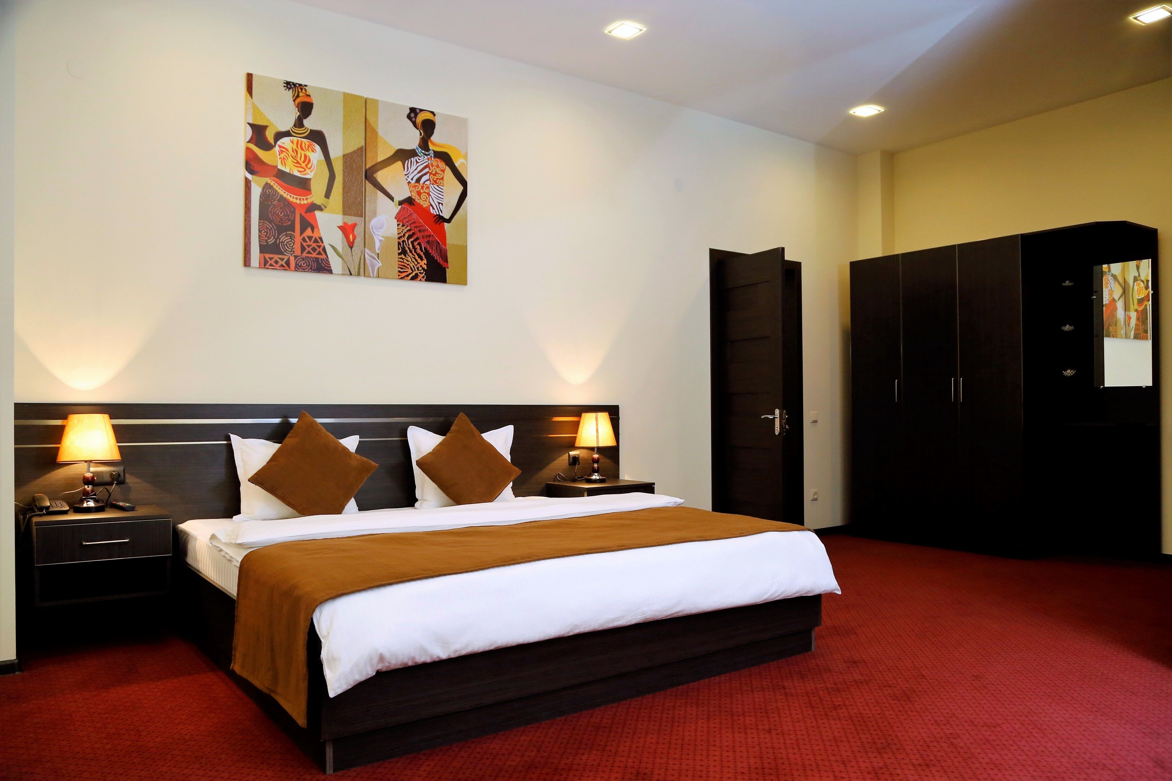 A hotel room with a large bed, two bedside tables with lamps, a wardrobe, and artwork on the walls.