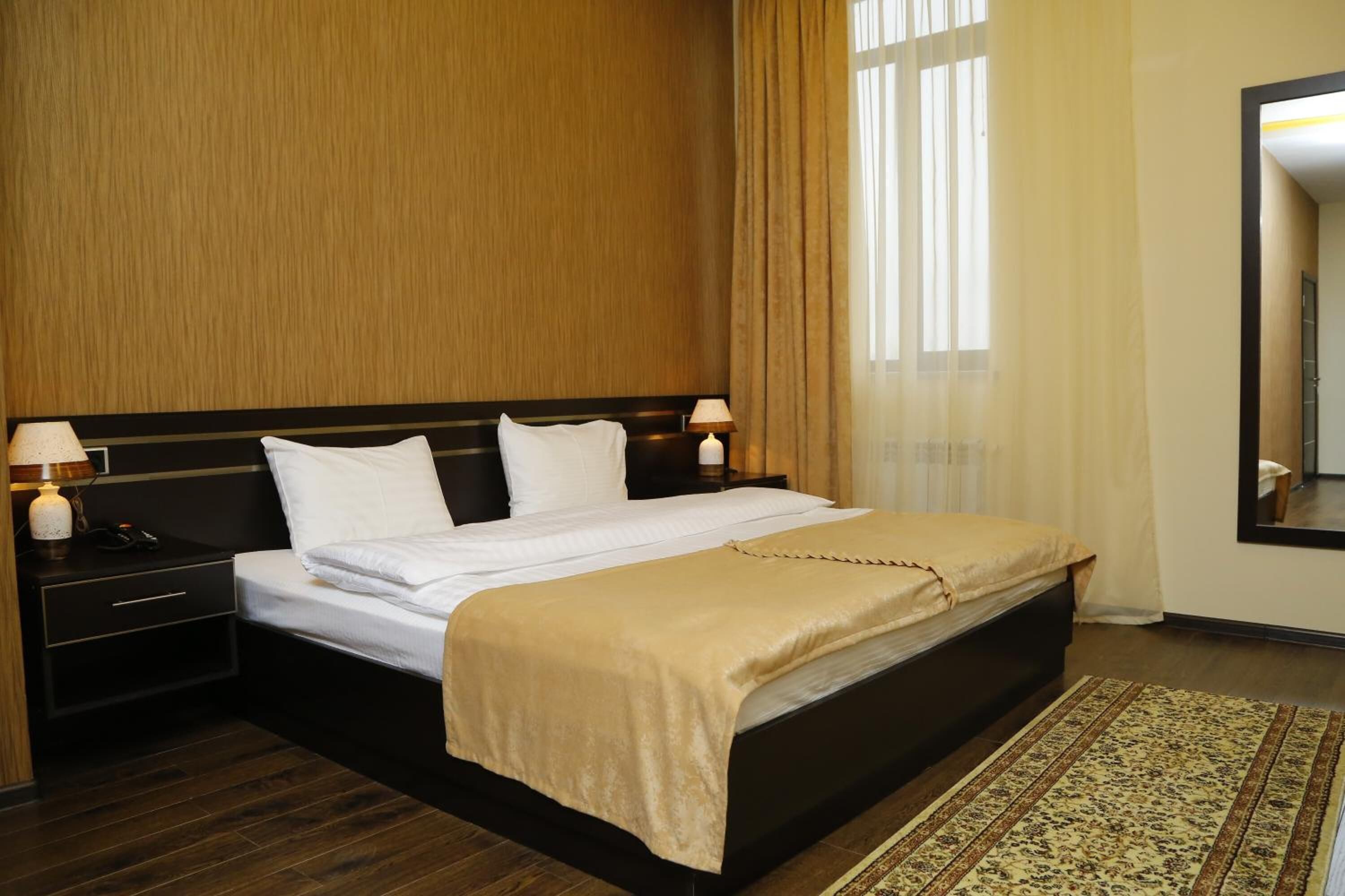 A hotel room with a large bed, wooden headboard, two bedside tables with lamps, a large mirror, and a patterned rug.