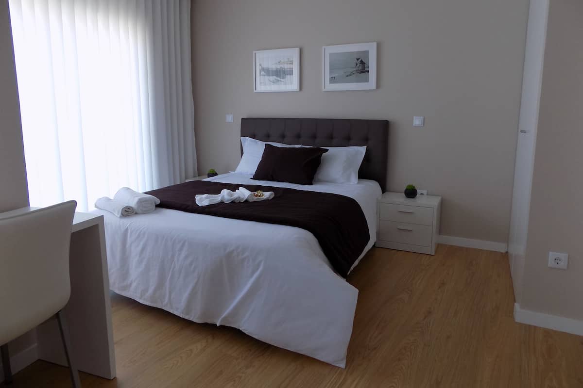A neatly made bed with white and brown bedding, a small bedside table with a plant, and two framed pictures on the wall.