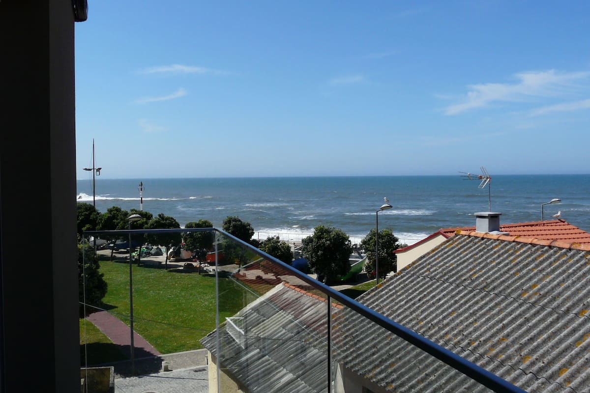 A coastal view from a balcony overlooking the sea, a lighthouse, and a street with parked cars and greenery.