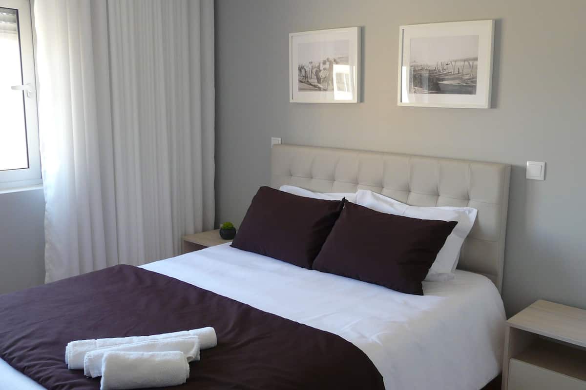 A neatly made bed with white and brown bedding, two framed pictures on the wall, and a window with curtains.