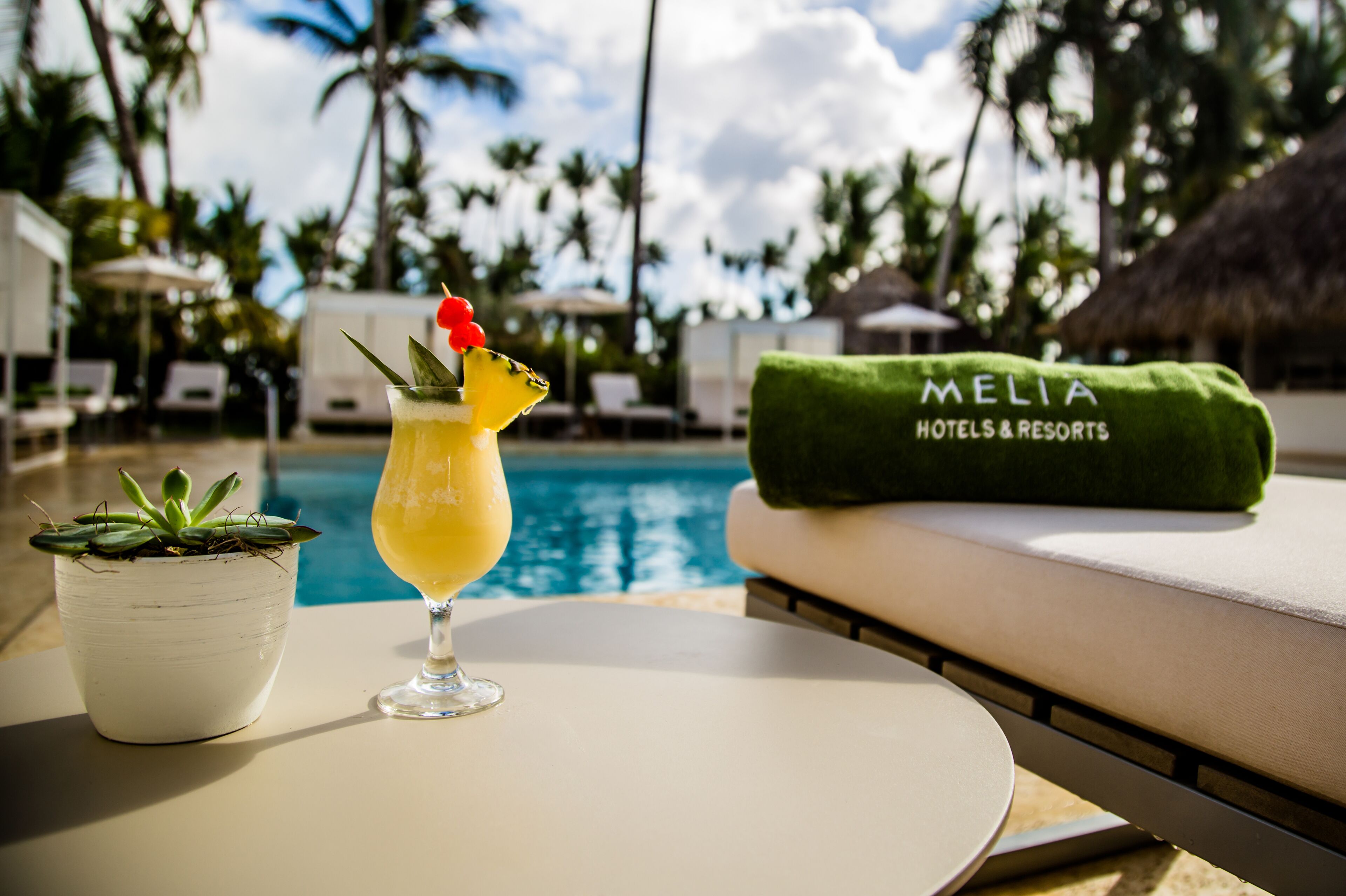 Area piscina con un drink, una pianta in vaso e un asciugamano con il marchio MELIA HOTELS & RESORTS.