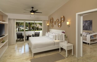 Un dormitorio amplio con una cama grande, ventilador de techo, televisión y un balcón con vistas a palmeras.