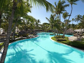 Relax by the resort's large swimming pool surrounded by lush palm trees and thatched-roof structures.