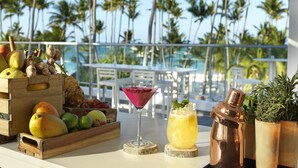 Un décor tropical avec une caisse en bois remplie de fruits, un verre à martini et un shaker à cocktail en cuivre, le tout sur un balcon donnant sur des palmiers et une piscine.