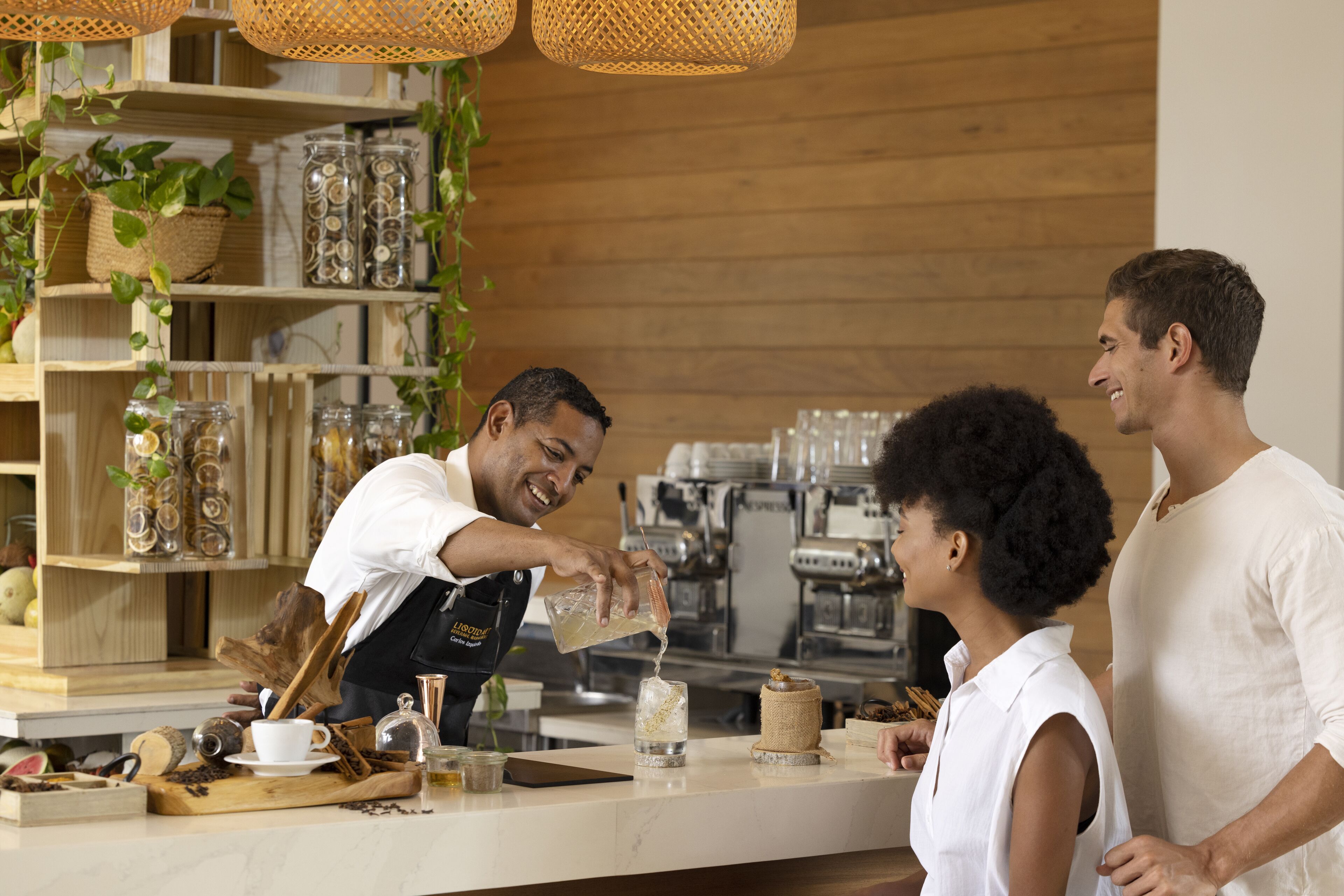 Un barista prépare une boisson au comptoir, avec des étagères remplies de bocaux et des plantes en arrière-plan.