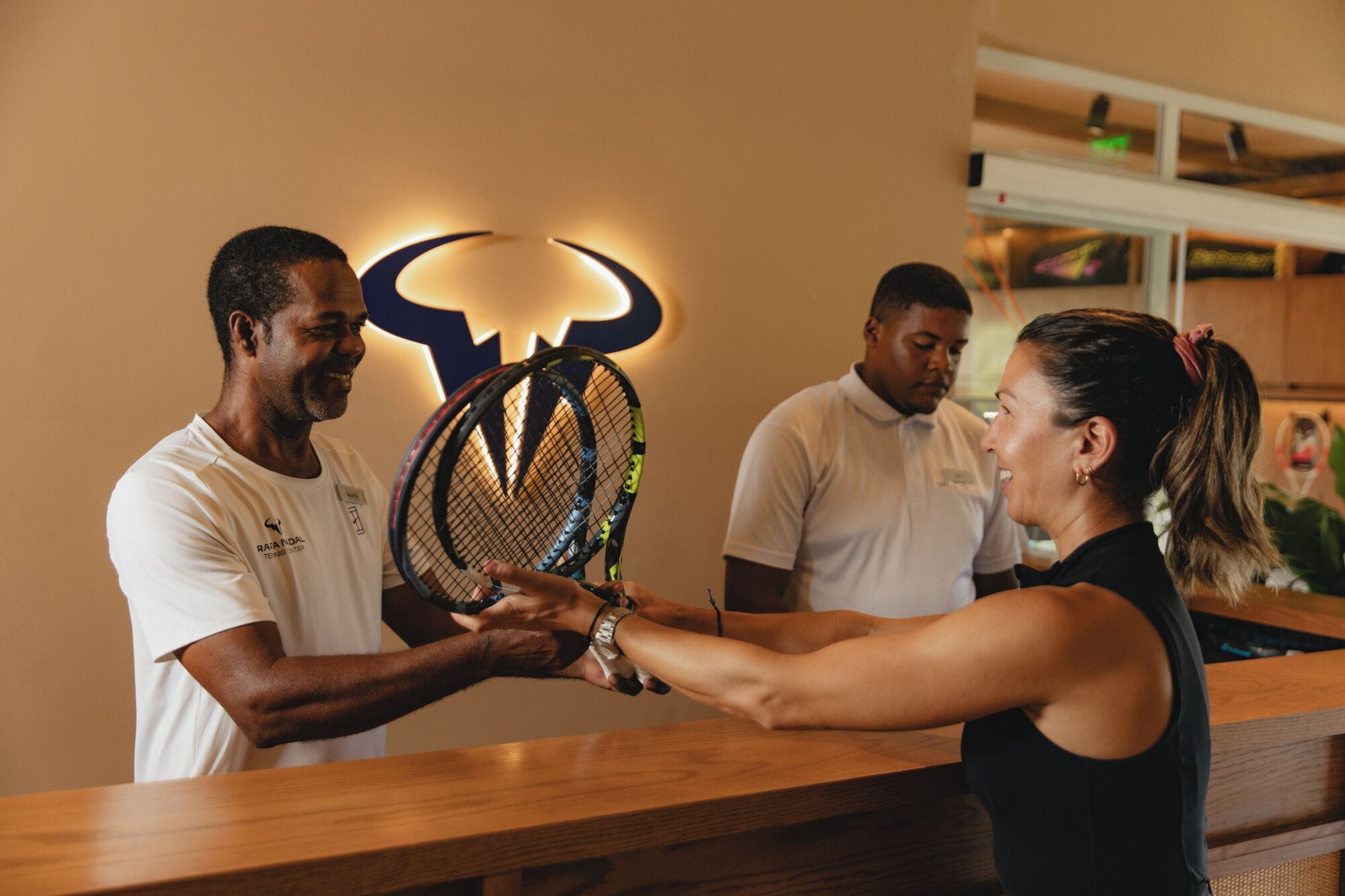 A reception area with a wooden counter, a logo on the wall, and a person holding a tennis racket.