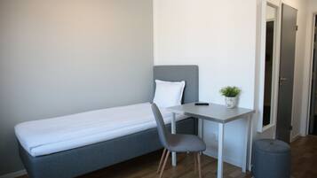 A compact room with a single bed, a small desk, a chair, and a potted plant.