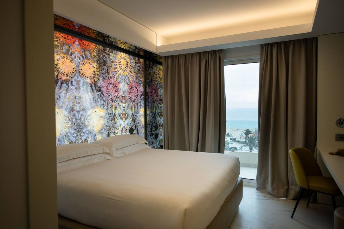 Een hotelkamer met een groot bed, uitzicht op zee en een kleurrijke muurschildering.