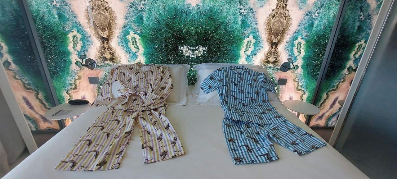 Una cama con dos vestidos estampados encima, una mesita de noche con una lámpara, y una pared con un mural de temática natural.