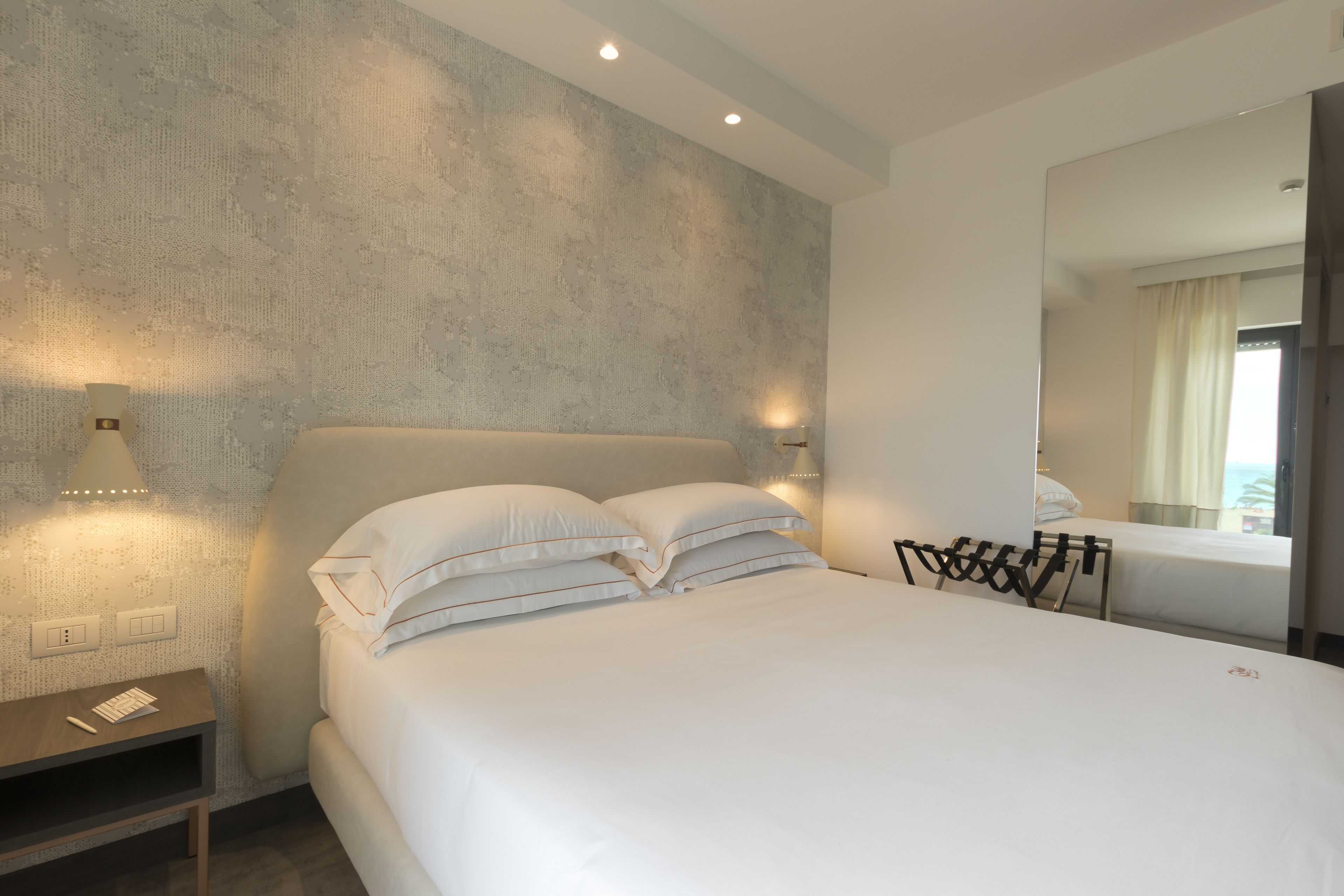 A modern hotel room with a large bed, bedside lamps, a nightstand, and a mirror reflecting the room.