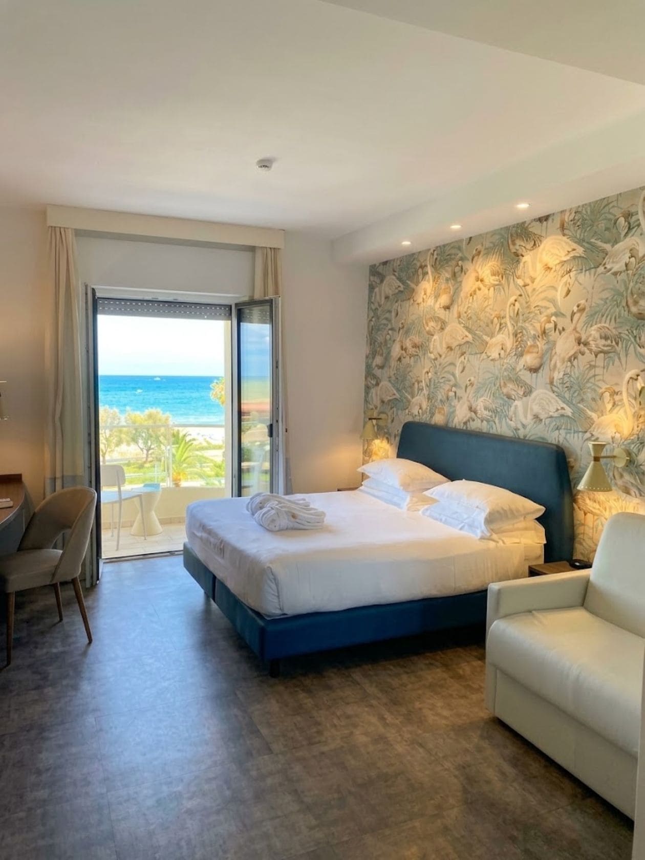 A hotel room with a bed, a desk, a chair, a view of the ocean, and a mural on the wall.
