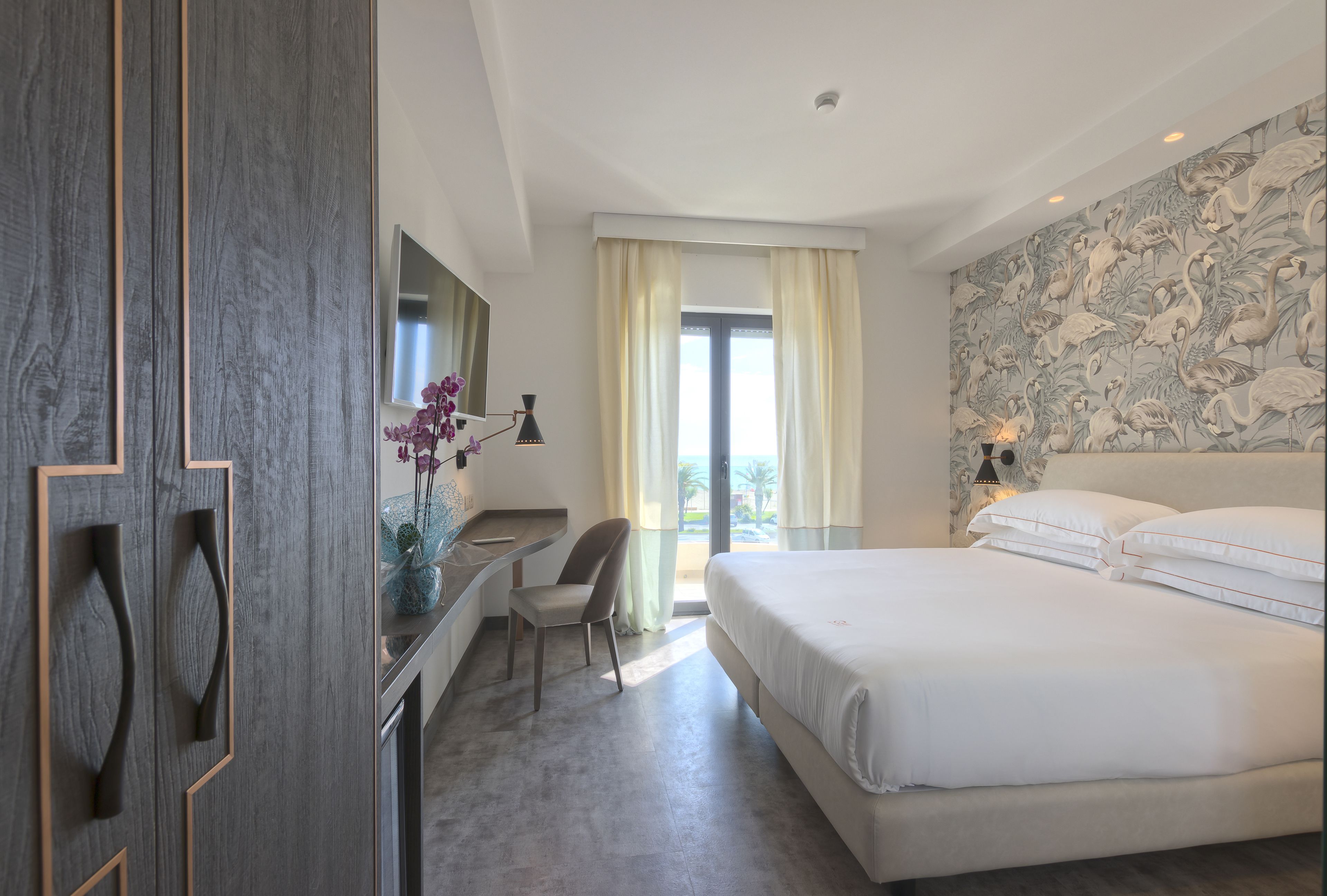 Une chambre d’hôtel avec un lit, un bureau, une chaise, un miroir et une fenêtre avec des rideaux.