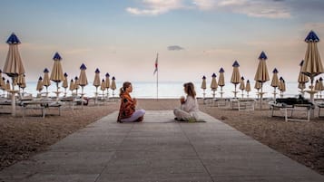 Two individuals sitting on a wooden pathway at a beach with beach umbrellas and lounge chairs in the background.