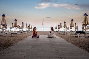 Two individuals sitting on a wooden pathway at a beach with beach umbrellas and lounge chairs in the background.