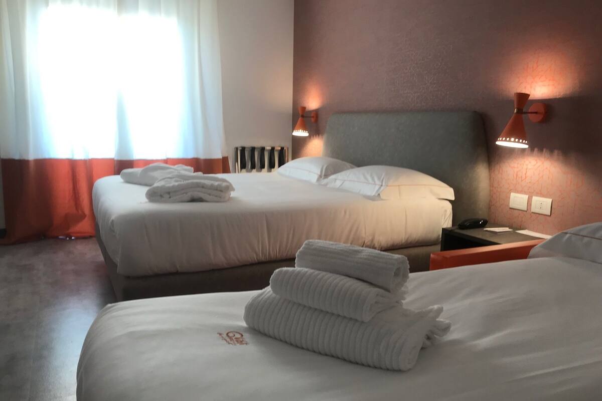 A hotel room with two beds, a bedside table with a phone and papers, a wall-mounted lamp, and a window with curtains.
