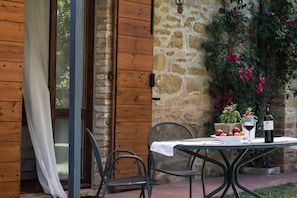 A wooden exterior wall with a stone wall, a window with a curtain, and a table with a bowl of fruit and a bottle of wine. - B&B Villa San Bartolomeo (Montepulciano)