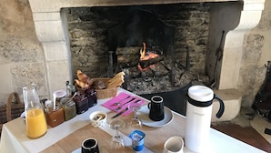 Een open haard met gloeiende blokken hout, een tafel met ontbijtartikelen, en diverse pompoenen op de schoorsteenmantel.