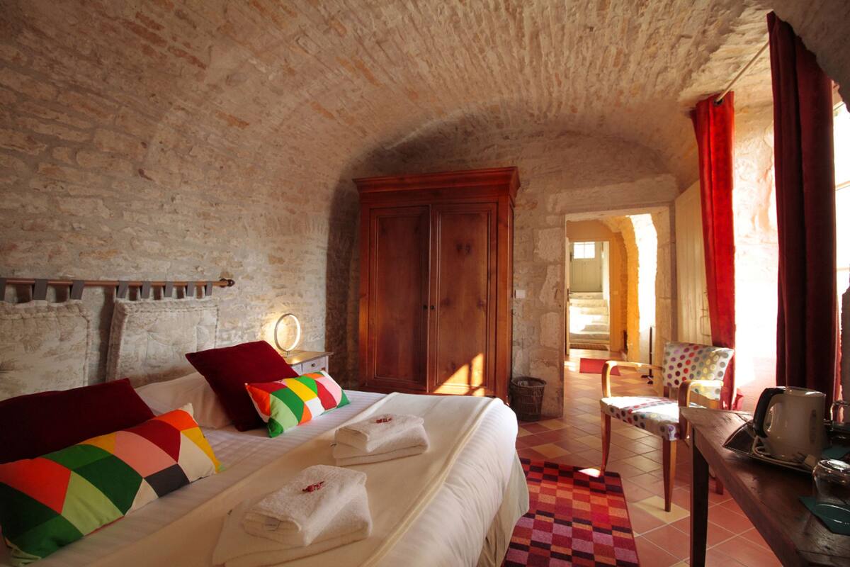 A bedroom with a large bed, a wooden wardrobe, a chair, and a table with a kettle.