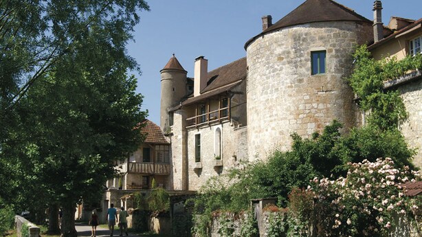 Een historisch gebouw van steen met een torentje en een balkon, omgeven door weelderige begroeiing en een pad.