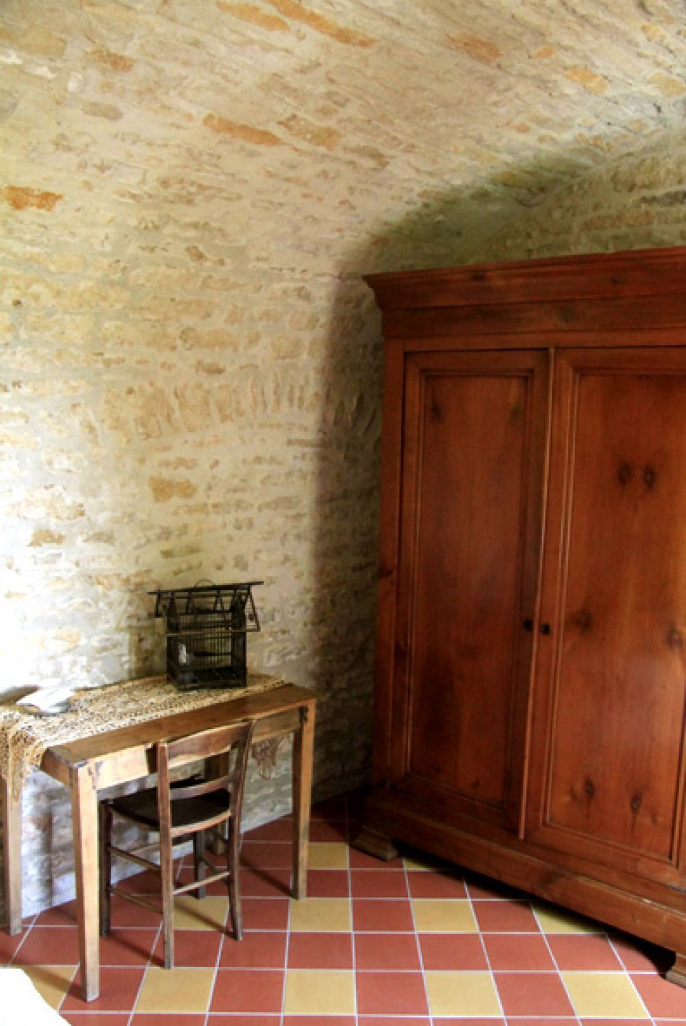 Une pièce avec un mur en pierre, une armoire en bois, une table en bois avec une chaise, et un sol carrelé.