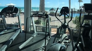 A gym with treadmills and exercise equipment, overlooking a beach with palm trees and a thatched umbrella. - Encanto Living 1101 -2BR by Kivoya (Puerto Peñasco)