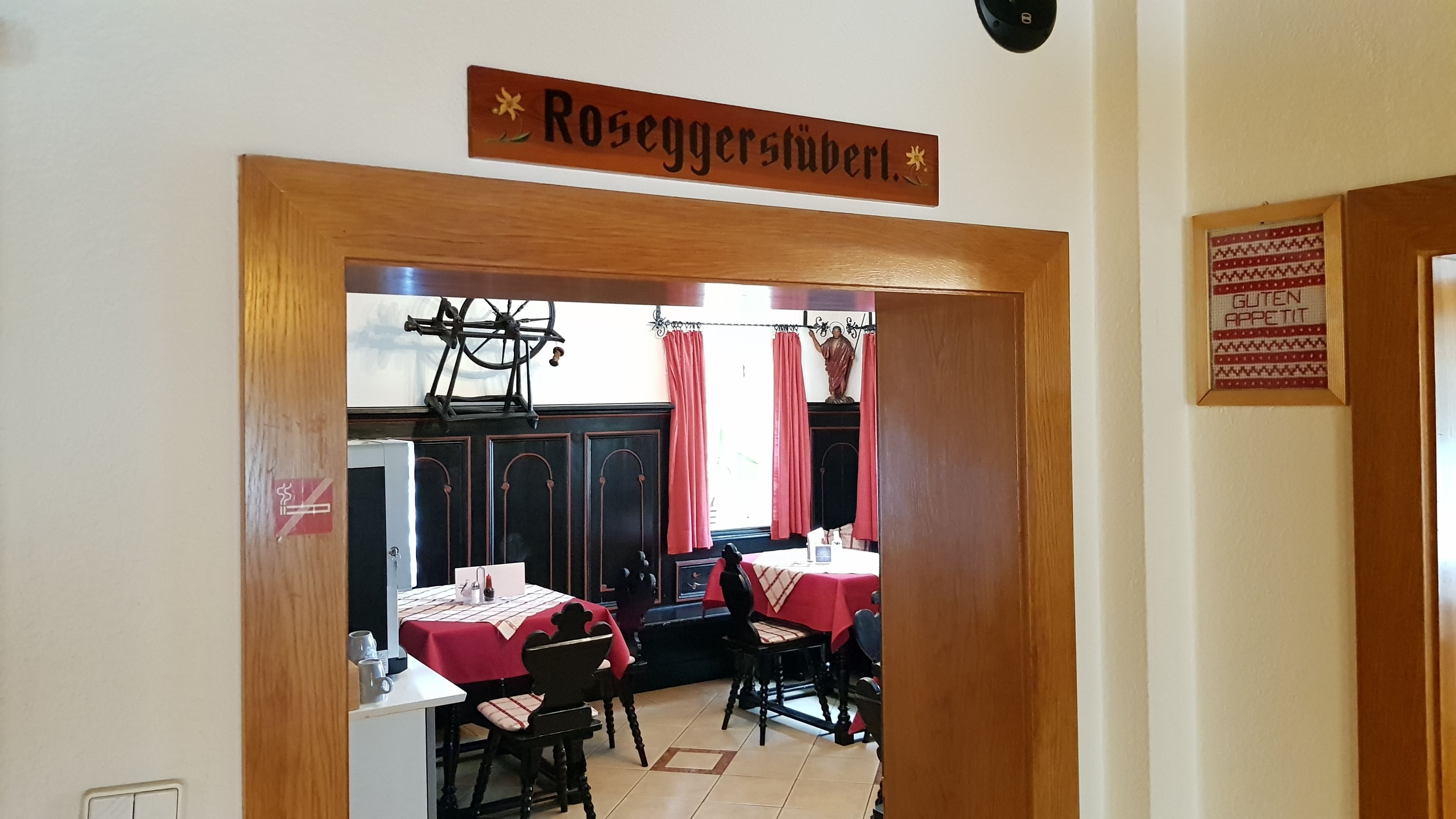 Een eetruimte met tafels en stoelen, een bord boven het hoofd met de tekst 'Roseggershütte', en een lijstje aan de muur met 'Eet smakelijk'.