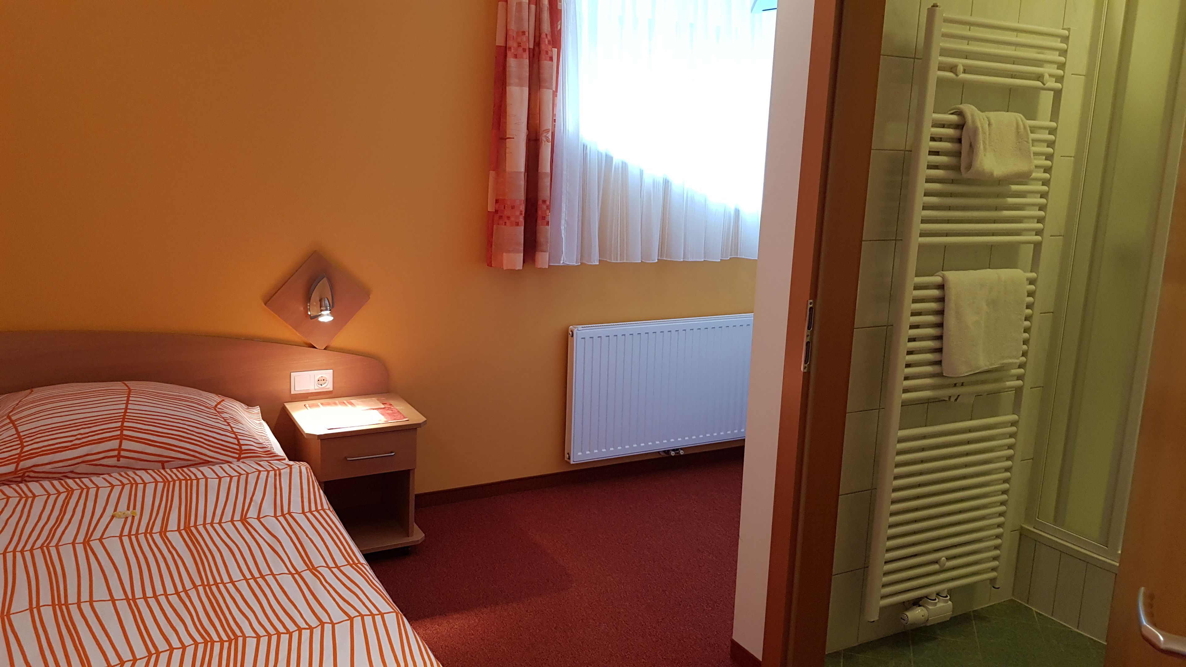 Ein Hotelzimmer mit Bett, Nachttisch, Lampe, Fenster mit Vorhängen, Heizkörper und Handtuchhalter.