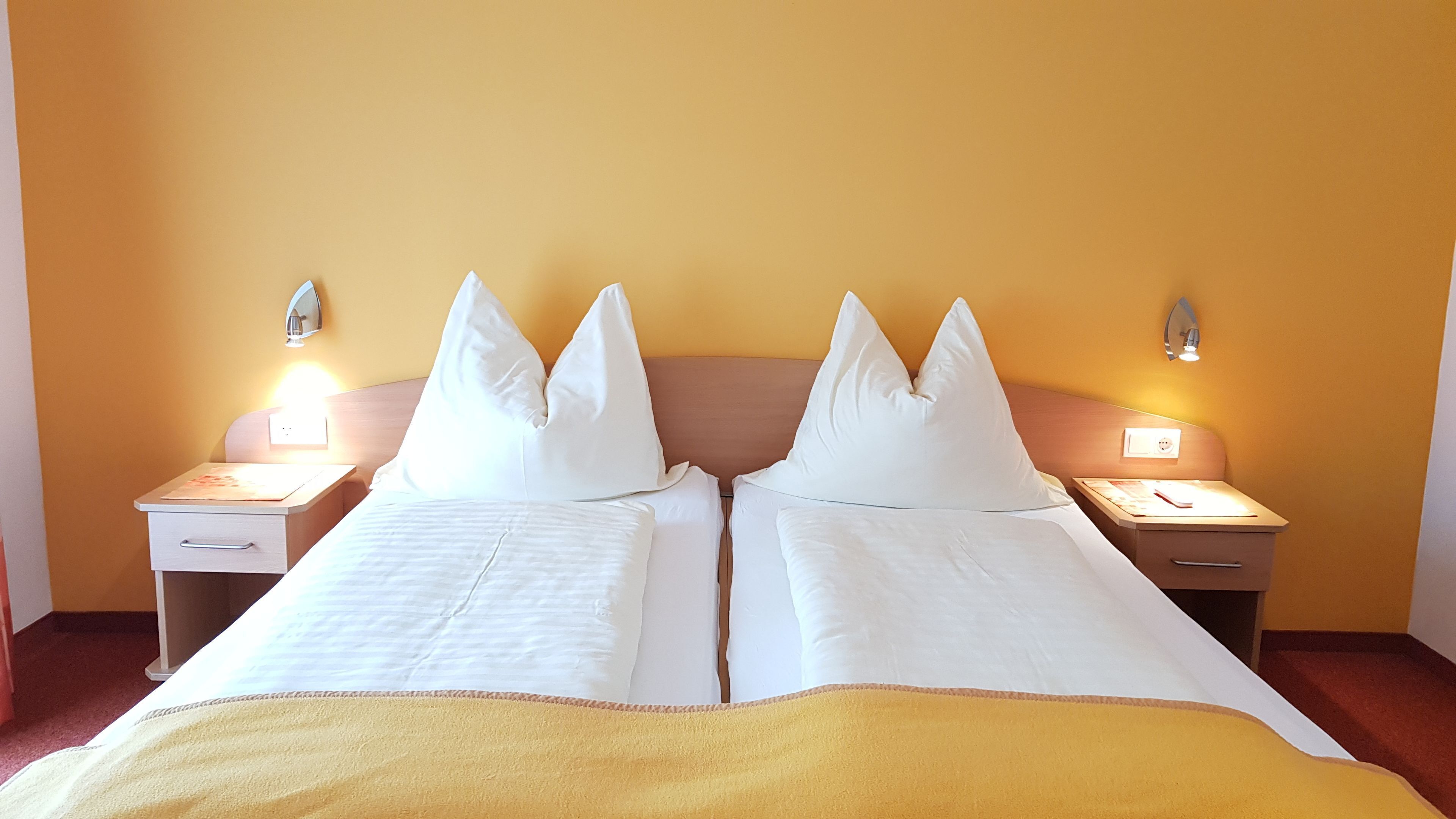 Un lit double avec du linge de lit blanc et un plaid jaune, deux tables de chevet avec des lampes, et un mur jaune uni.