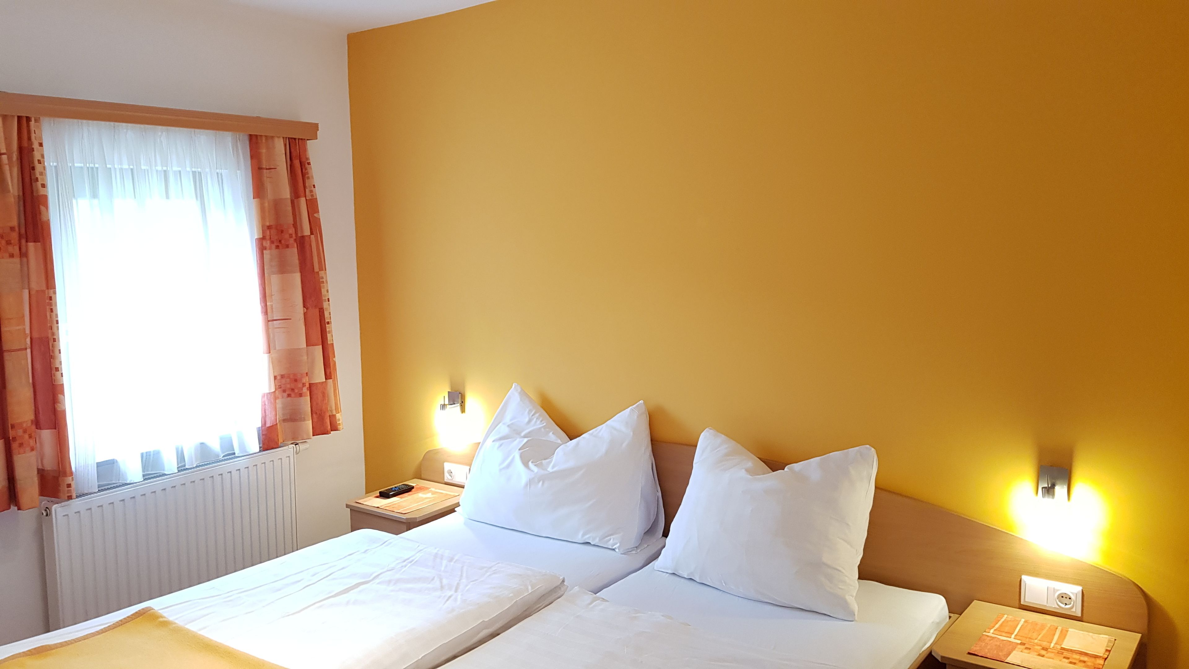 A hotel room with a bed, two bedside tables, a nightstand, a window with curtains, and a wall-mounted light fixture.
