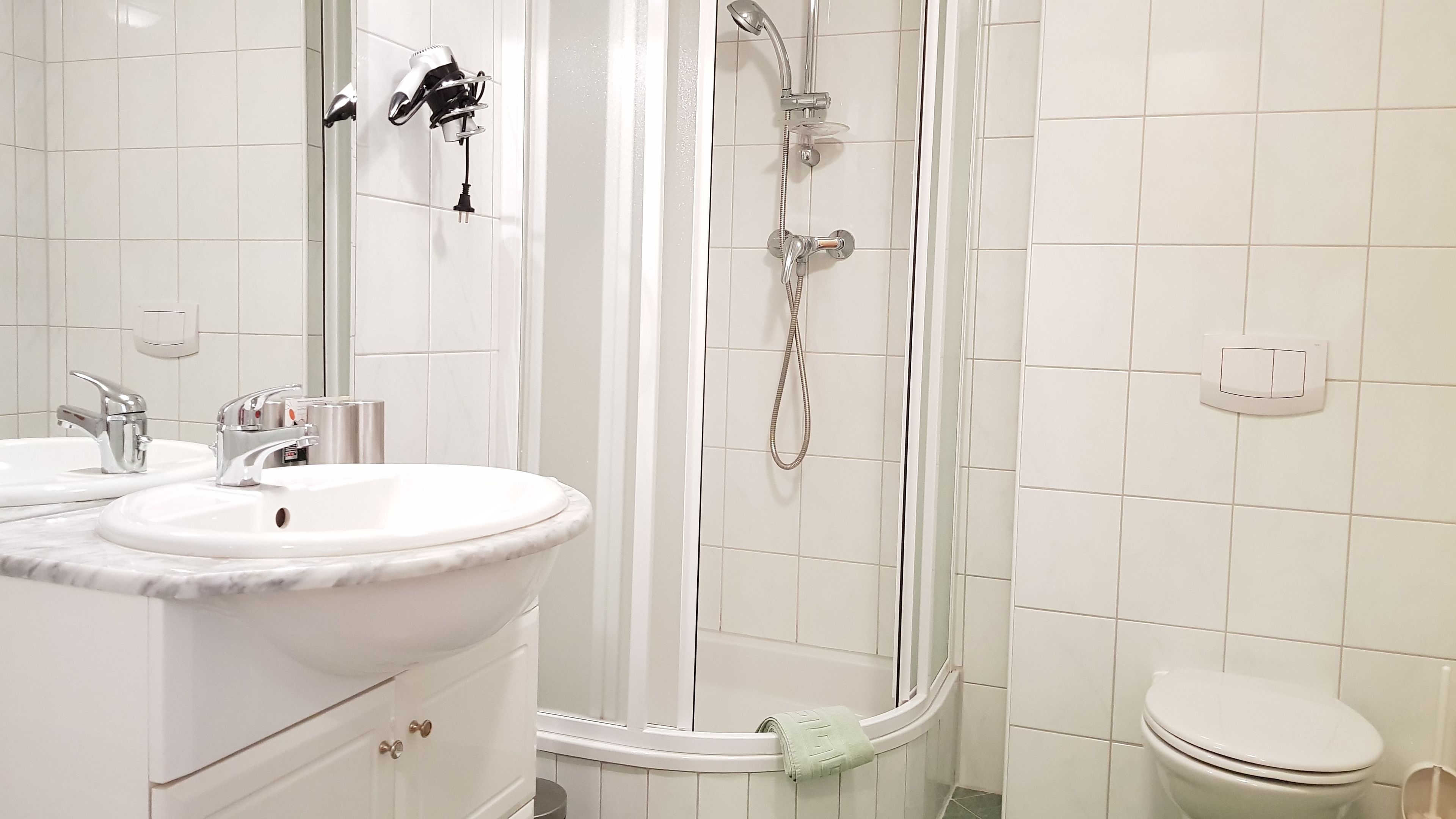 Ein Badezimmer mit einem weißen Waschbecken, zwei Wasserhähnen, einem Spiegel, einer Dusche mit Duschbrause und einer Toilette.