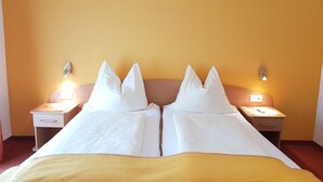Una cama doble con sábanas blancas y dos almohadas, una mesita de noche con lámpara, y un aplique de pared.