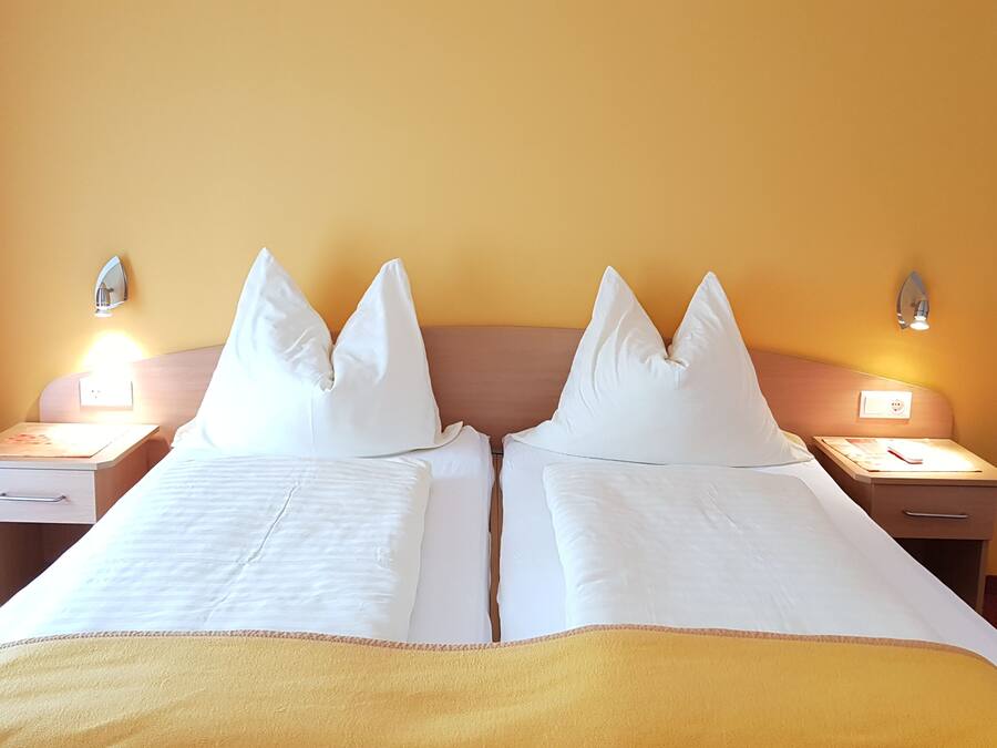 A double bed with white linens and two pillows, a bedside table with a lamp, and a wall-mounted light fixture.