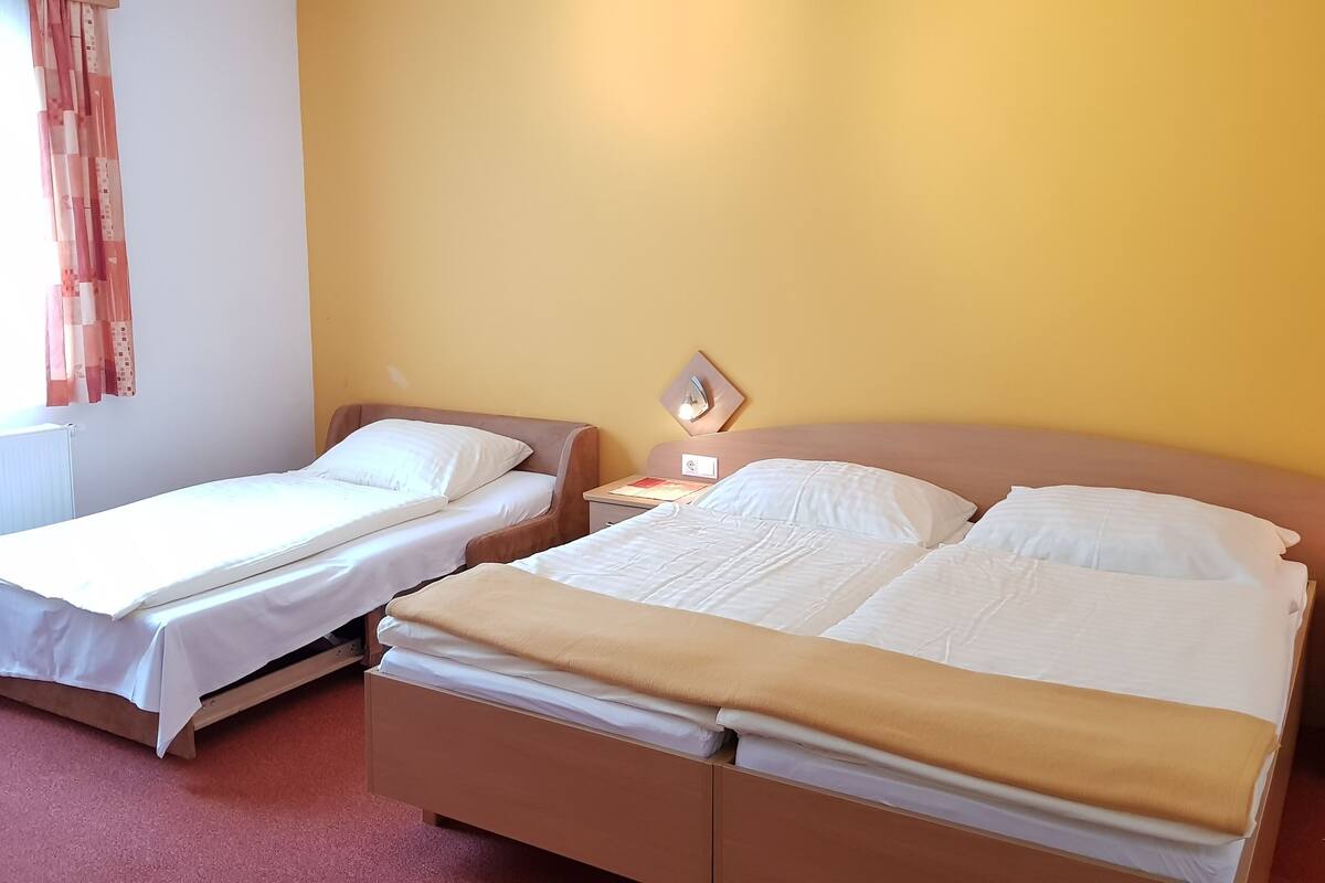 Une chambre d’hôtel avec deux lits simples et un lit double, une table de chevet avec une lampe et une fenêtre avec des rideaux.