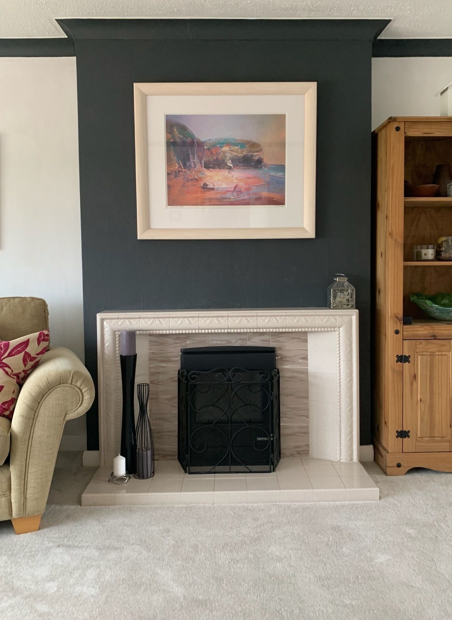 A living room with a fireplace, a framed painting above it, a beige armchair, and a wooden cabinet.