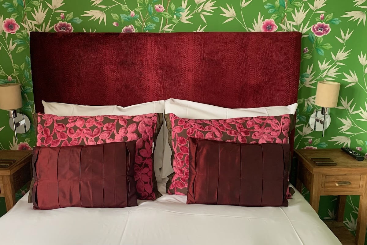 Een bed met een rood hoofdeinde, kussens met een bloemendessin en groene bloemenbehang op de achtergrond.