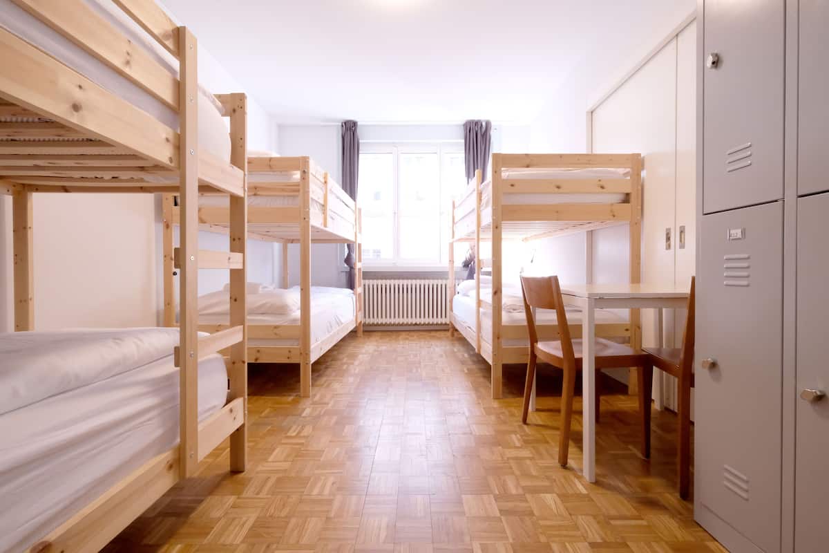 A dormitory room with bunk beds, a wooden desk, a chair, and a radiator.